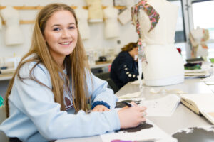 A student sits at a table with pen in hand. She smiles at the camera. A mannequin is on the table next to her.