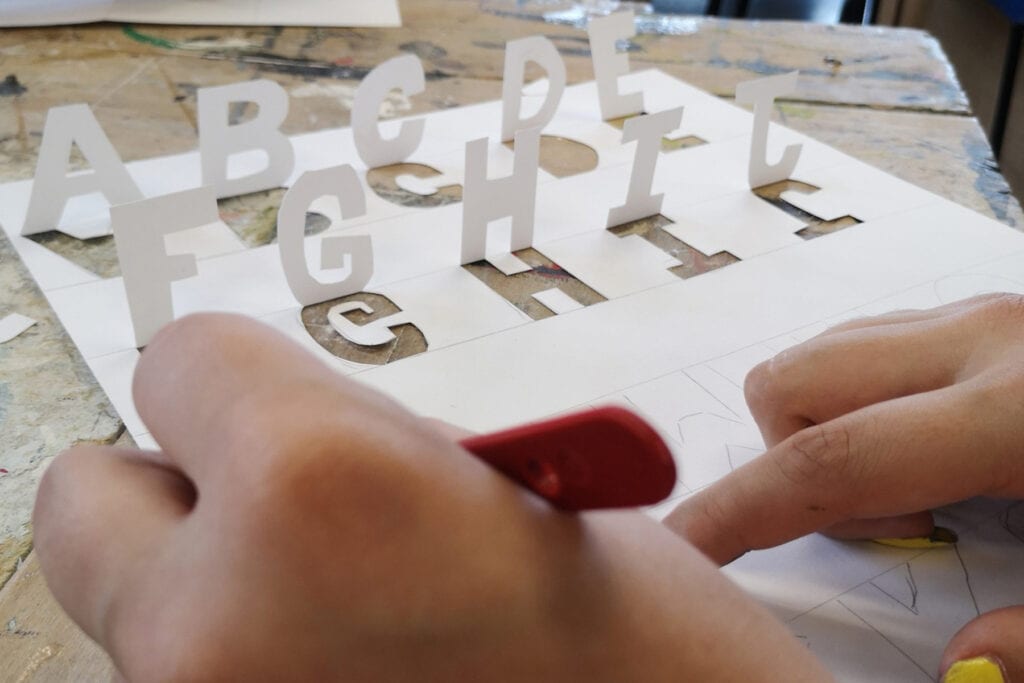 A close-up image of letters that have been carved from a piece of card. The letters are stood up vertically. Hands hold a scalpel.