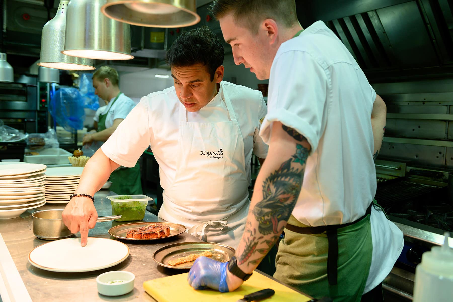 A photo of Paul Ainsworth with a chef in a working kitchen. They both look to two dishes and a clean plate.
