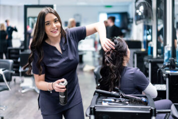 A hair and beauty student uses hairspray on another student within a professional salon environment.