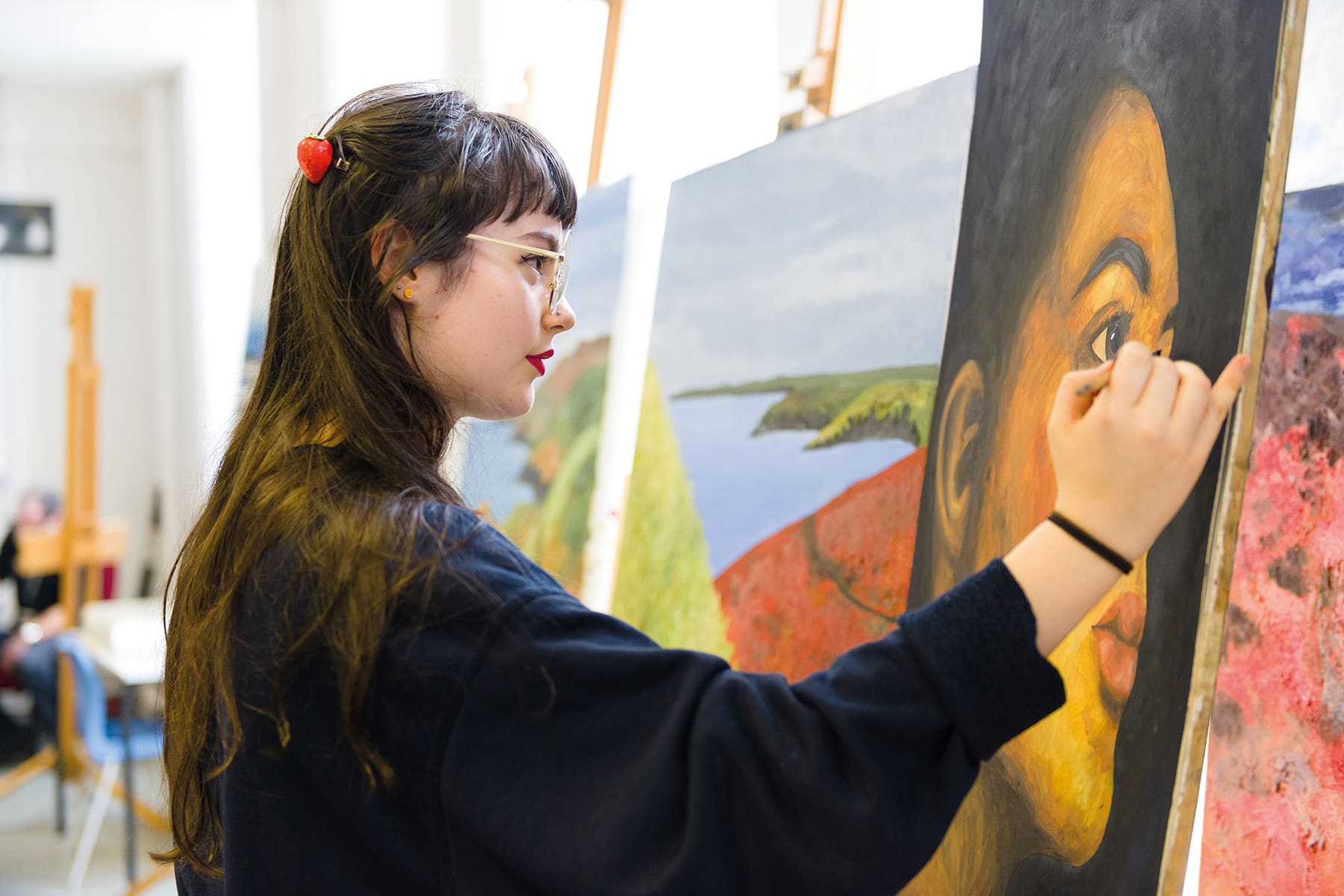 A student is painting on canvas on an easel. There are other paintings surrounding them.