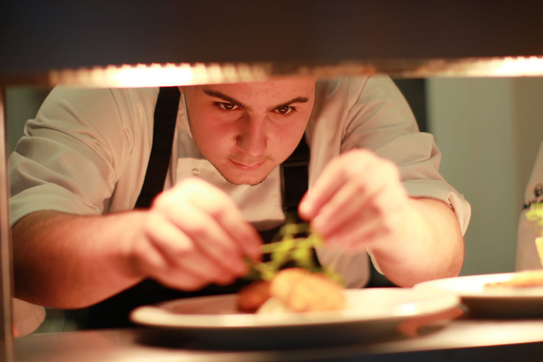 A chef leans down under heat lamps and puts the final dressing on a dish.