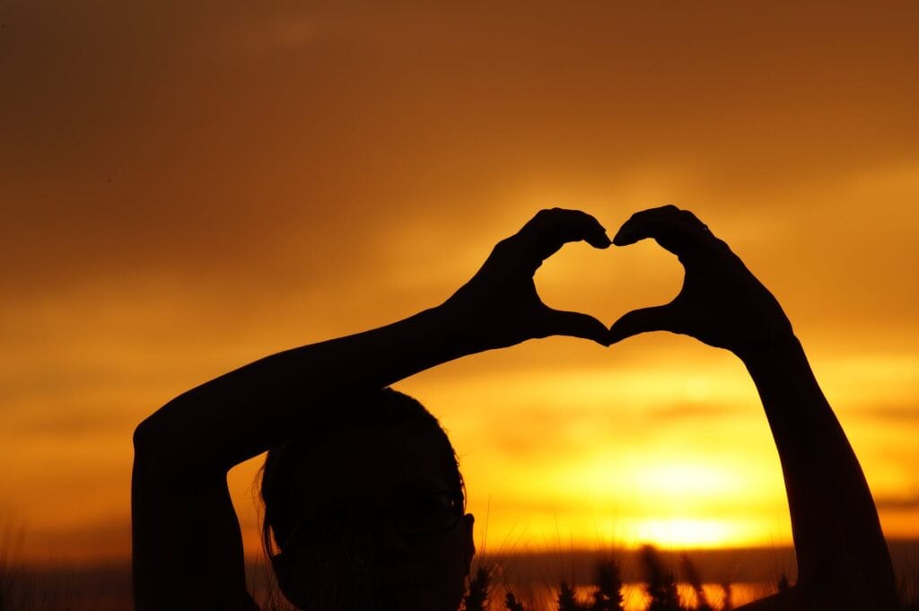 A silhouette of a person makes a heart sign with their hands above their head. There is a sunset behind them.