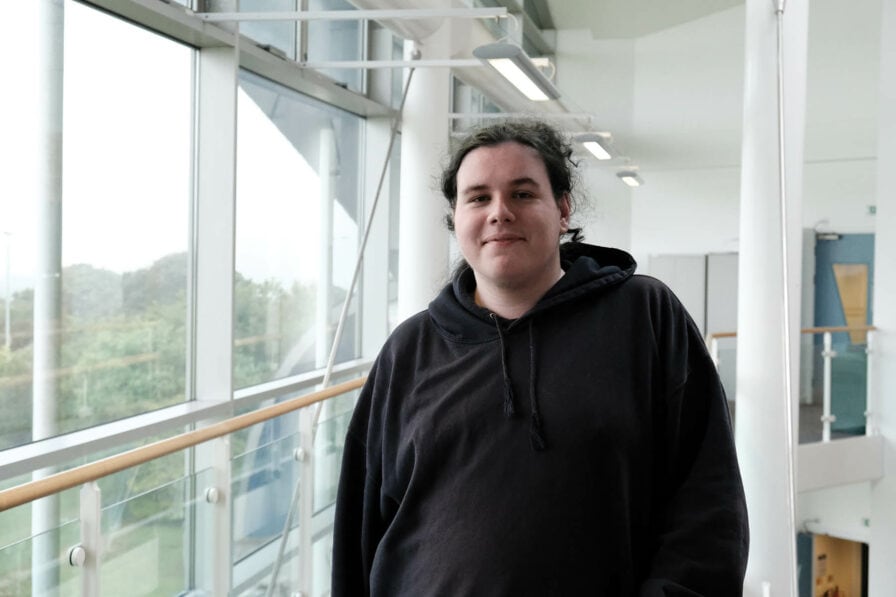 Aaron O'Brien Student Aaron stands in a corridor with lots of windows in a dark jumper.