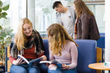 Two adult students sit and look through books together as two student chat behind them.