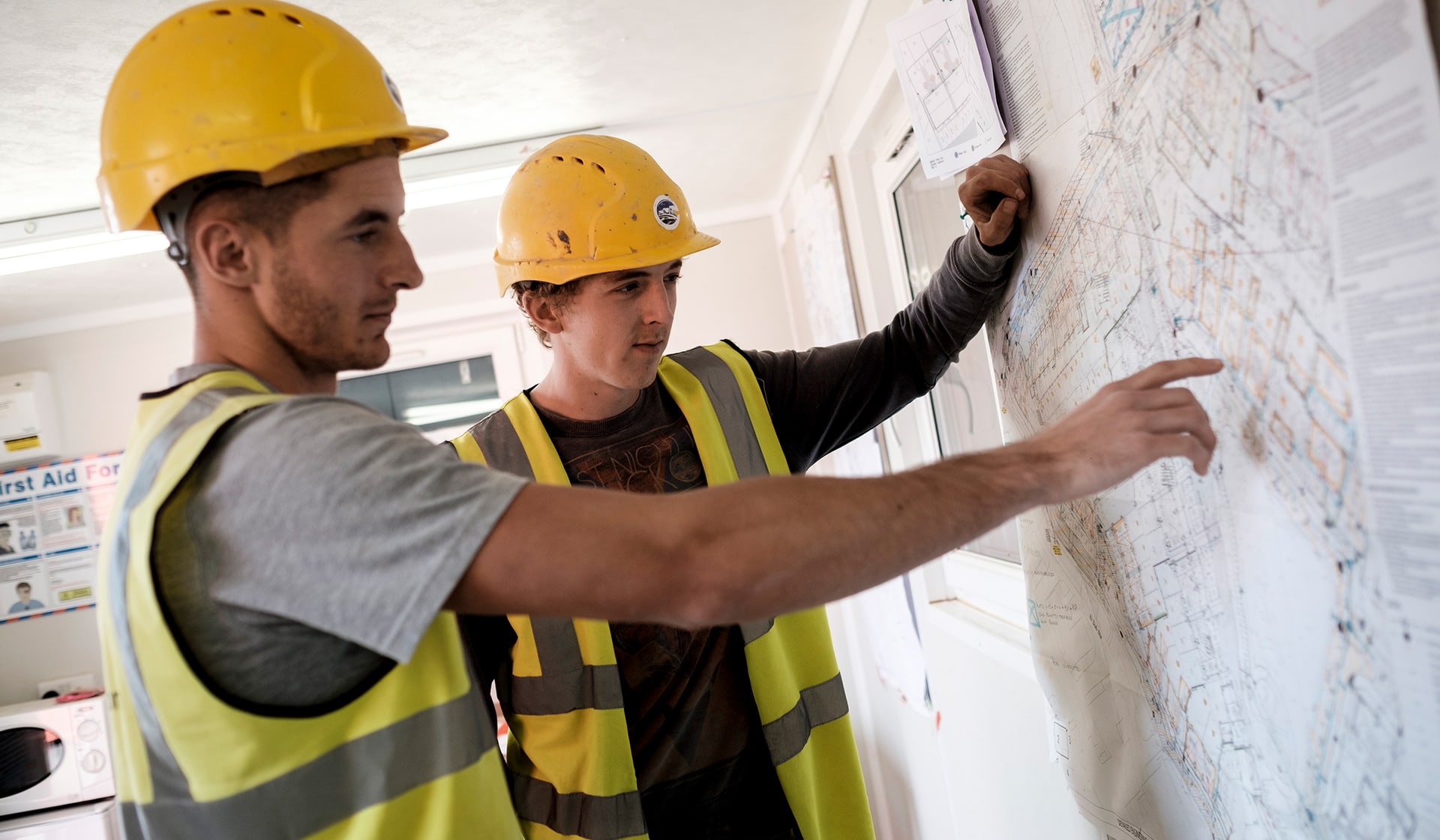Two construction workers wearing hi-vis jackets and hard hats look at a whiteboard together.