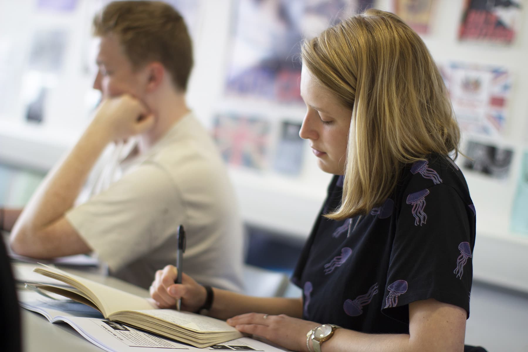 Two students sit at a classroom table with notepads and pens.