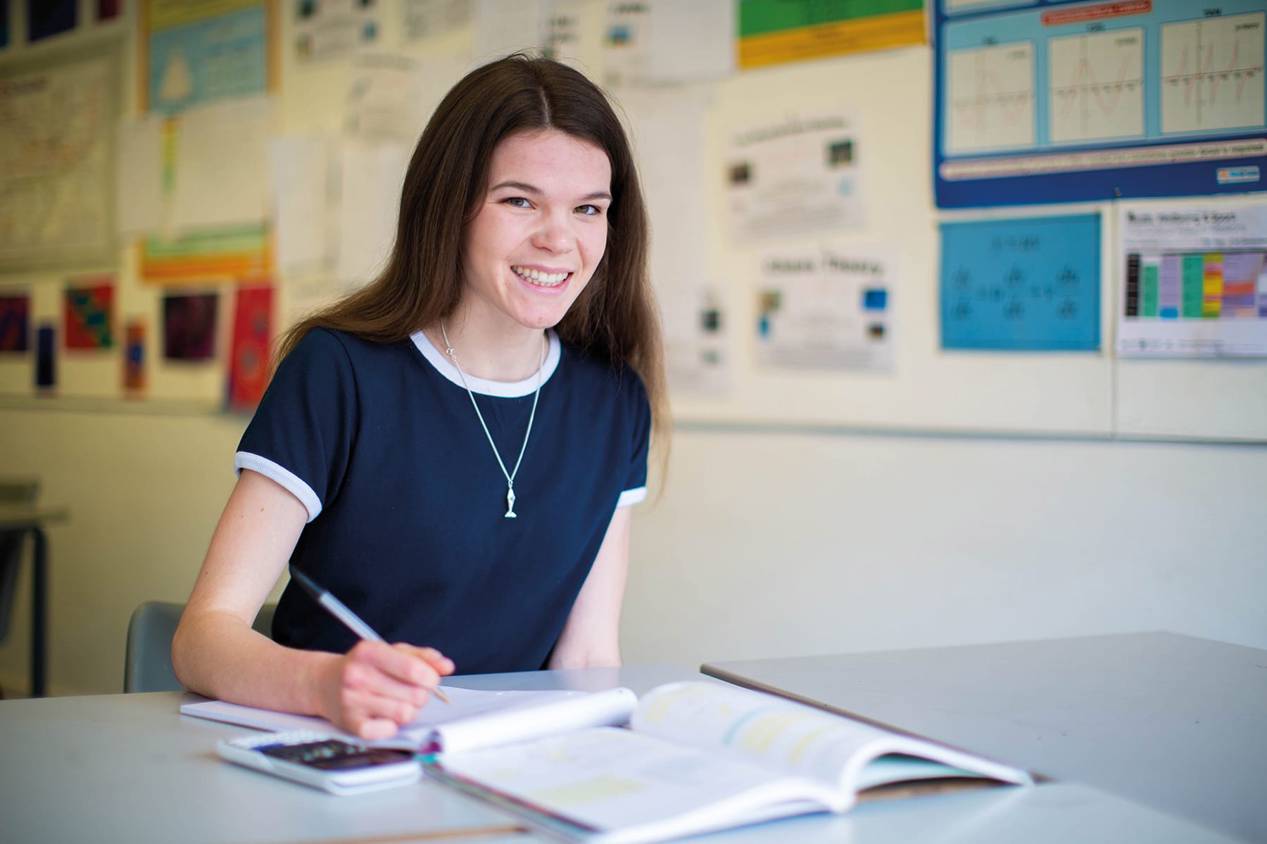 A mathematics student sits at a desk with pen and paper smiling at the camera.