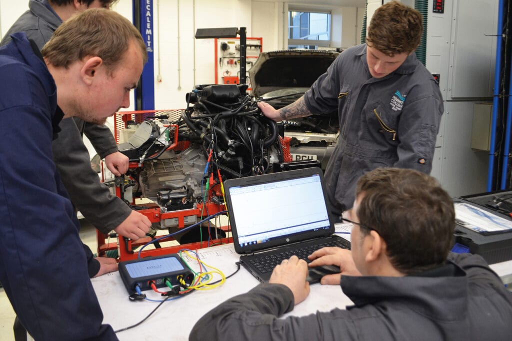 Students and staff look at a laptop within an automotive workshop.