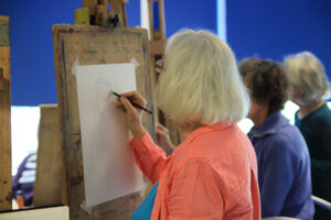 The camera looks over the shoulder of a woman who is sketching onto a piece of paper on an easel.