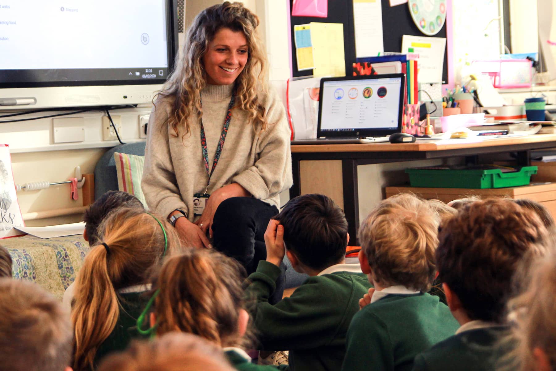 An Education student is teacher training, sits smiling at the front of a class of young primary school children who are all sat on the ground facing her.