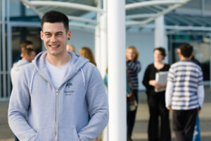 A young man looks at the camera smiling with his hands in the pockets of his hoodie. There are other people stood chatting behind him.