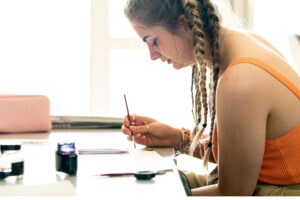An art student sits at a table with paint brush in hand, painting black swirl designs on white paper.
