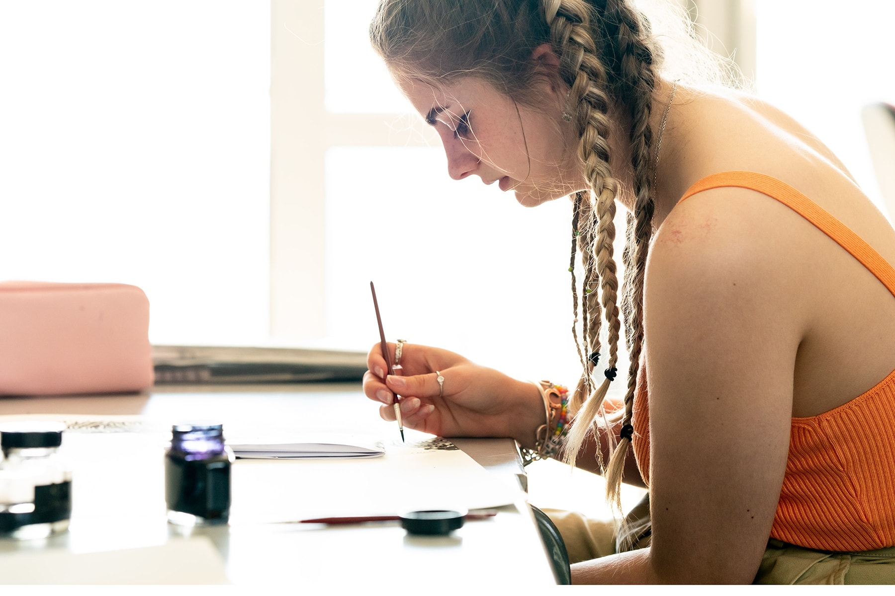 An art student sits at a table with paint brush in hand, painting black swirl designs on white paper.
