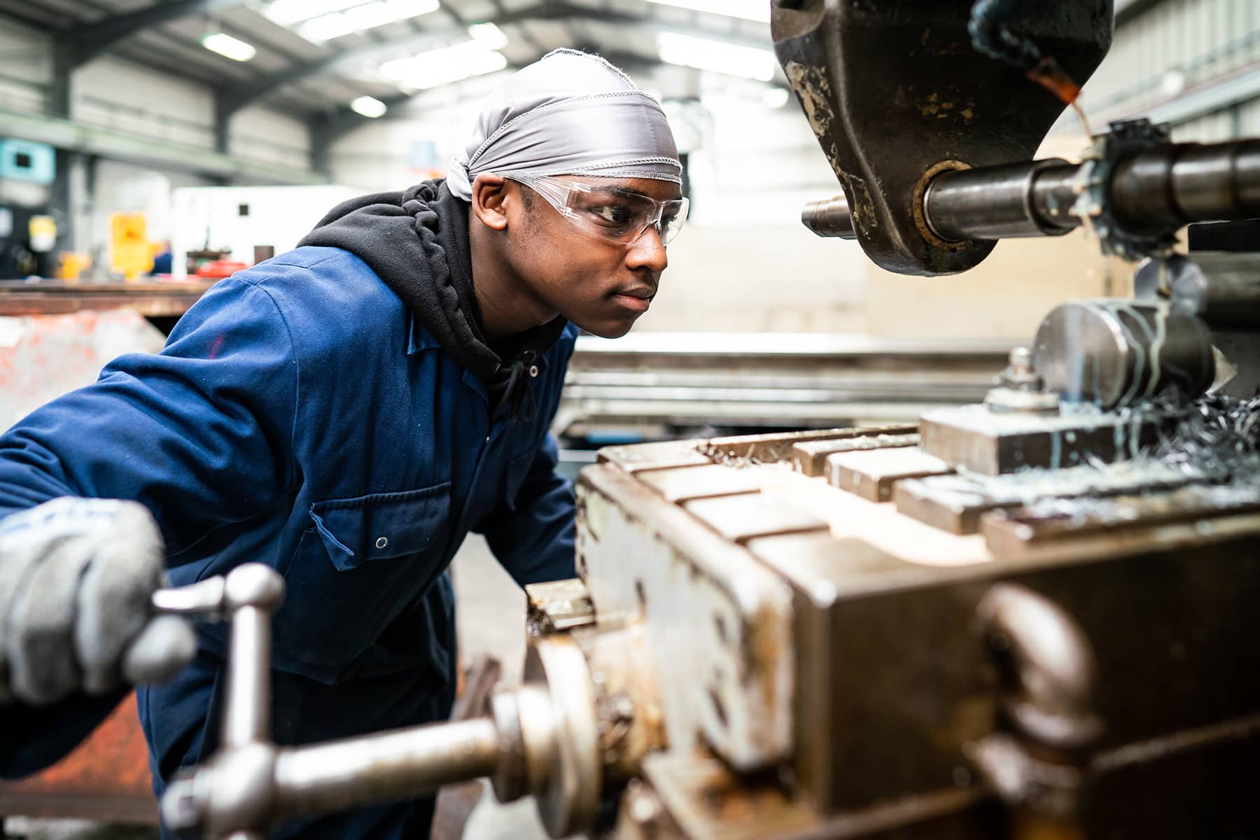 A young man wearing overalls and protective eyewear leans in close to large engineering equipment.