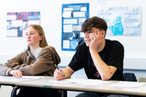 Two students sit at a classroom table looking towards the front of the class. One student has their hand to their chin and a pen in their other hand.