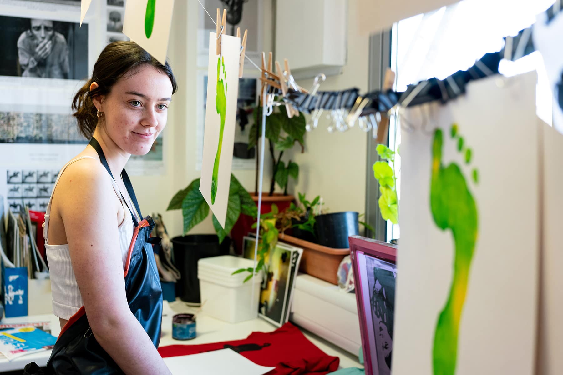 An Art & Design student is dressed in an apron and looking at art work hanging up to dry.