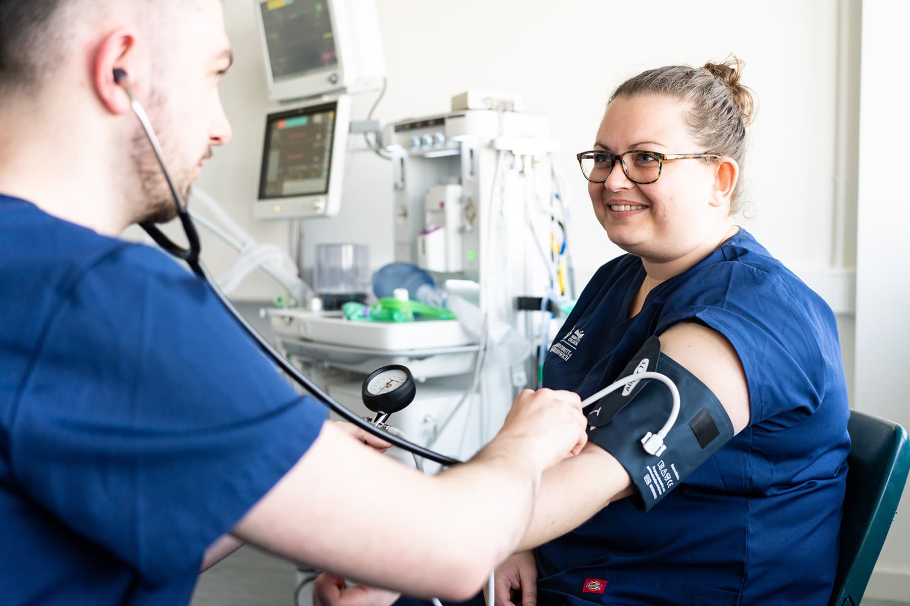 Two students dressed in scrubs take each others' blood pressure in a health and social care environment.