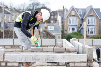 A young man wearing a high vis and hard hat lays bricks in a construction setting.