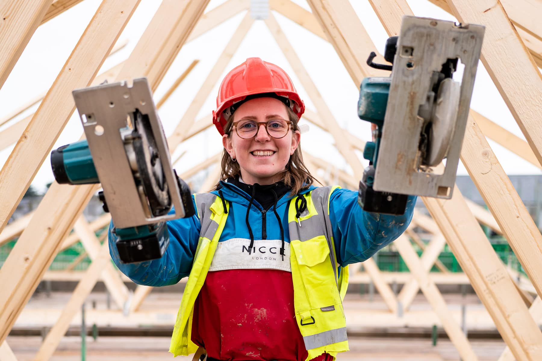 A construction apprentice lifts tools smiling at the camera in protective wear.