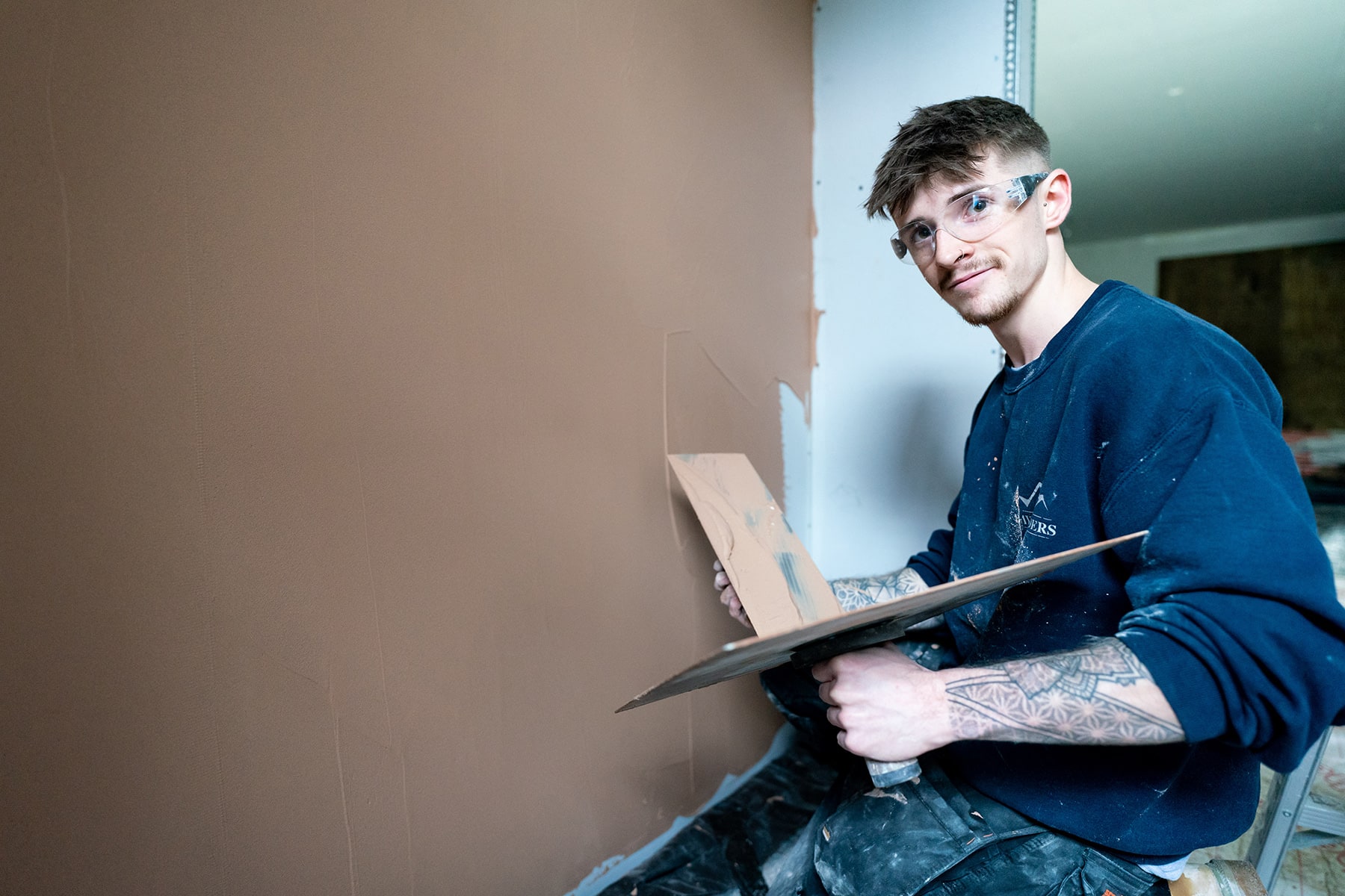 A construction student is plastering a wall looking at the camera.