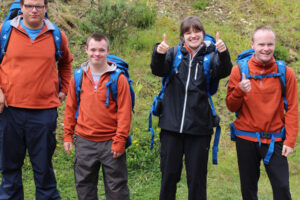A group of students wearing walking gear and backpacks look at the camera. One student does a double thumbs-up.