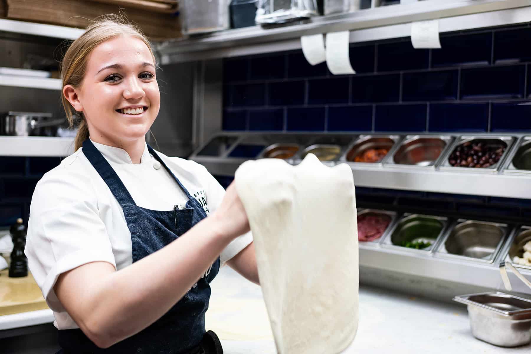 A young girl wearing an apron smiles at the camera as she stretches pizza dough in a professional kitchen.