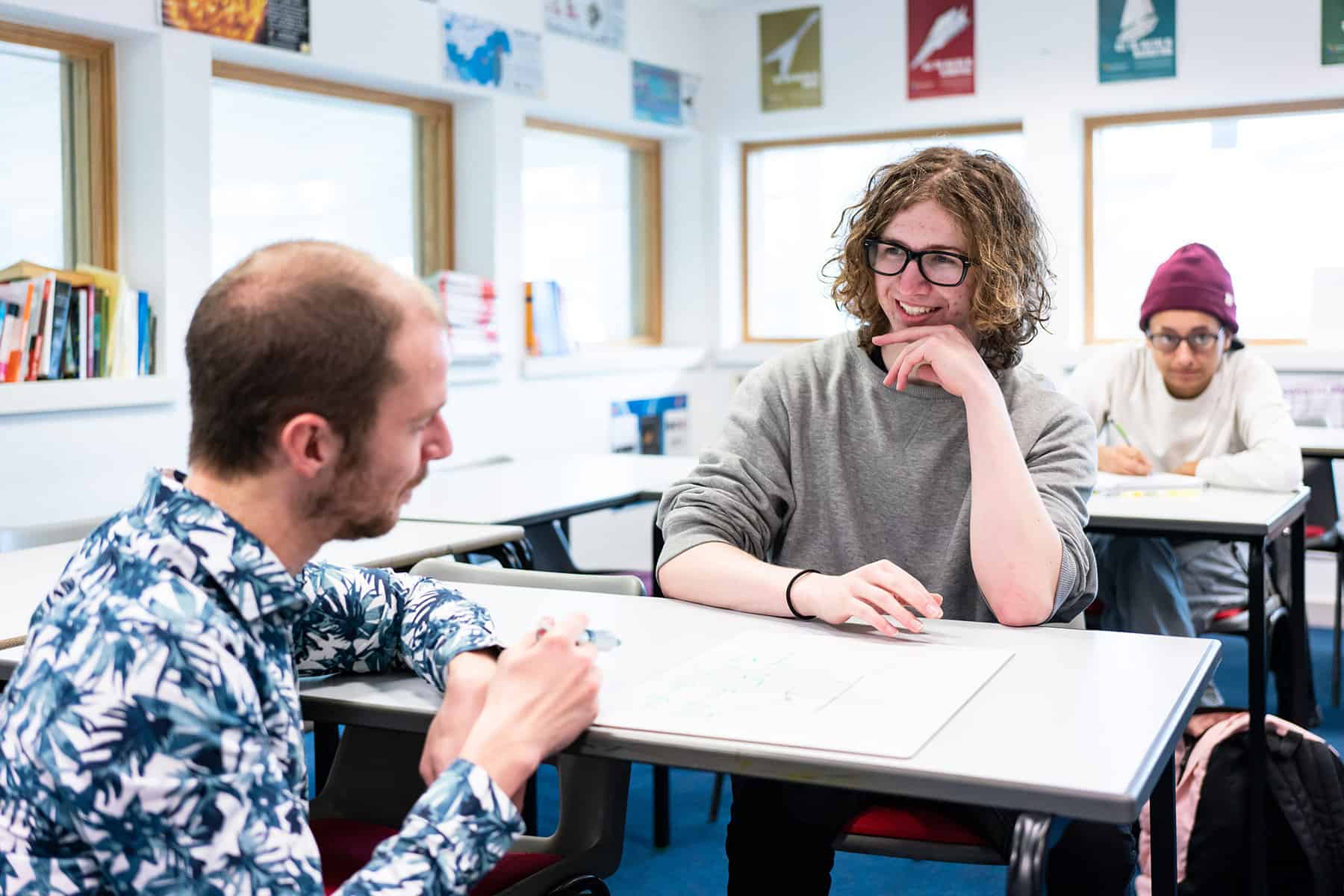 A tutor crouches down in front of a student who is sat at a desk. The student smiles.