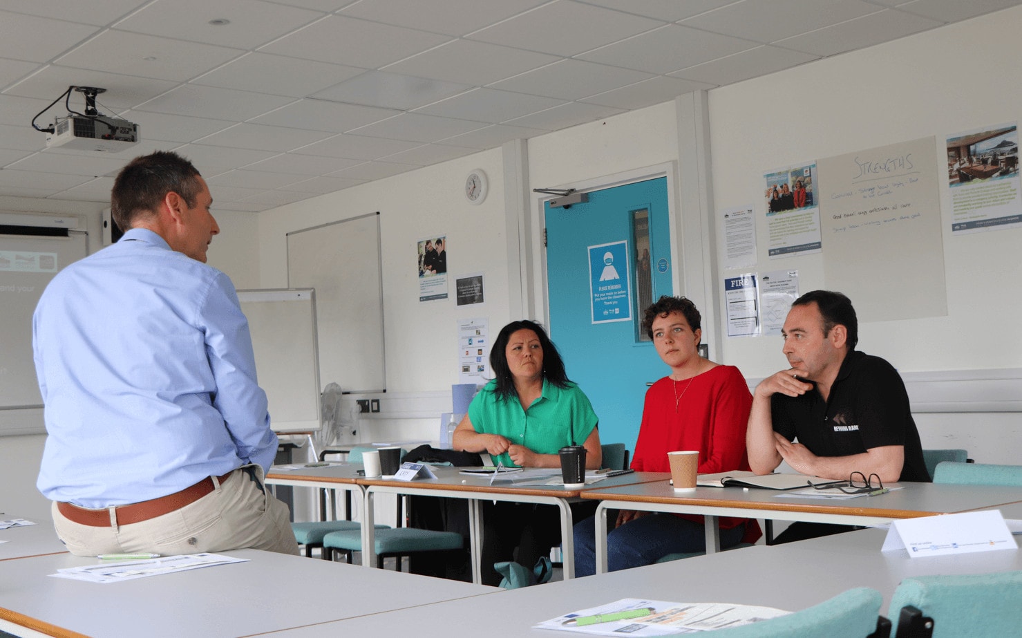 A photo of three adult students being taught in a classroom while the teacher sits on a desk in front of them.