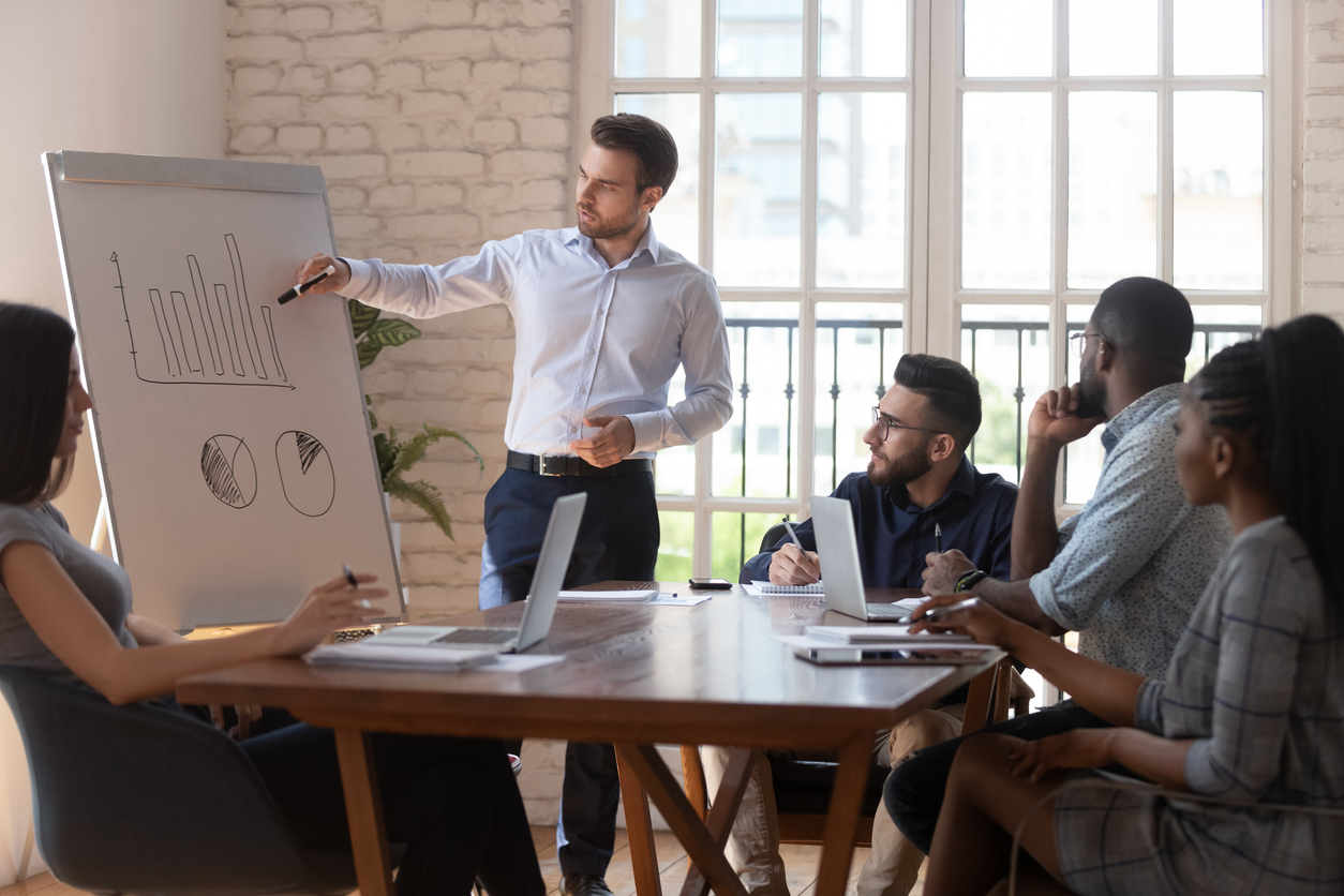 Confident male business trainer gives on flip chart presentation for diverse participants company staff, team leader explain graph strategy during corporate meeting seminar in modern light boardroom