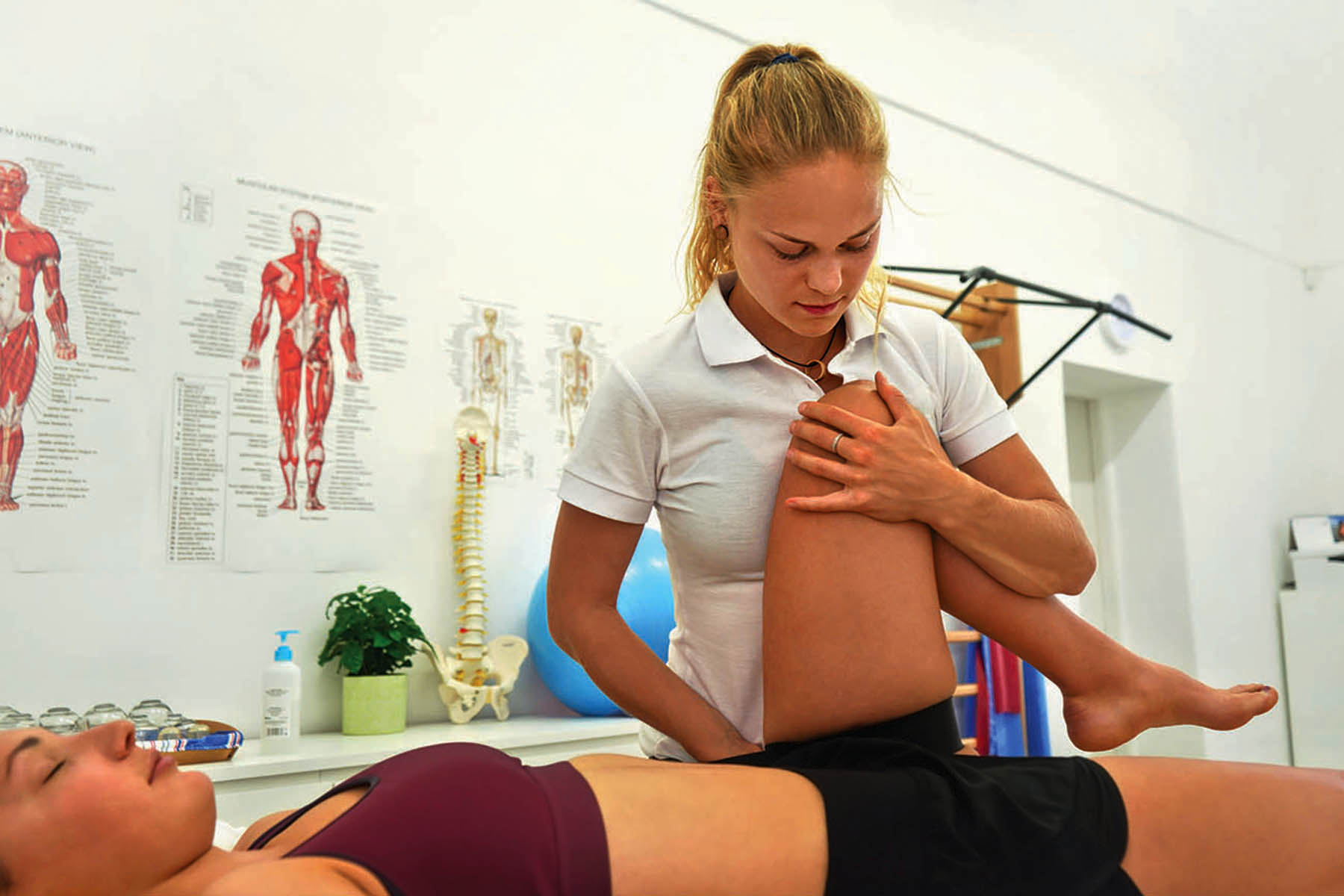 Young female physiotherapist exercising with her patient laying on massage table, flexing her knee muscles