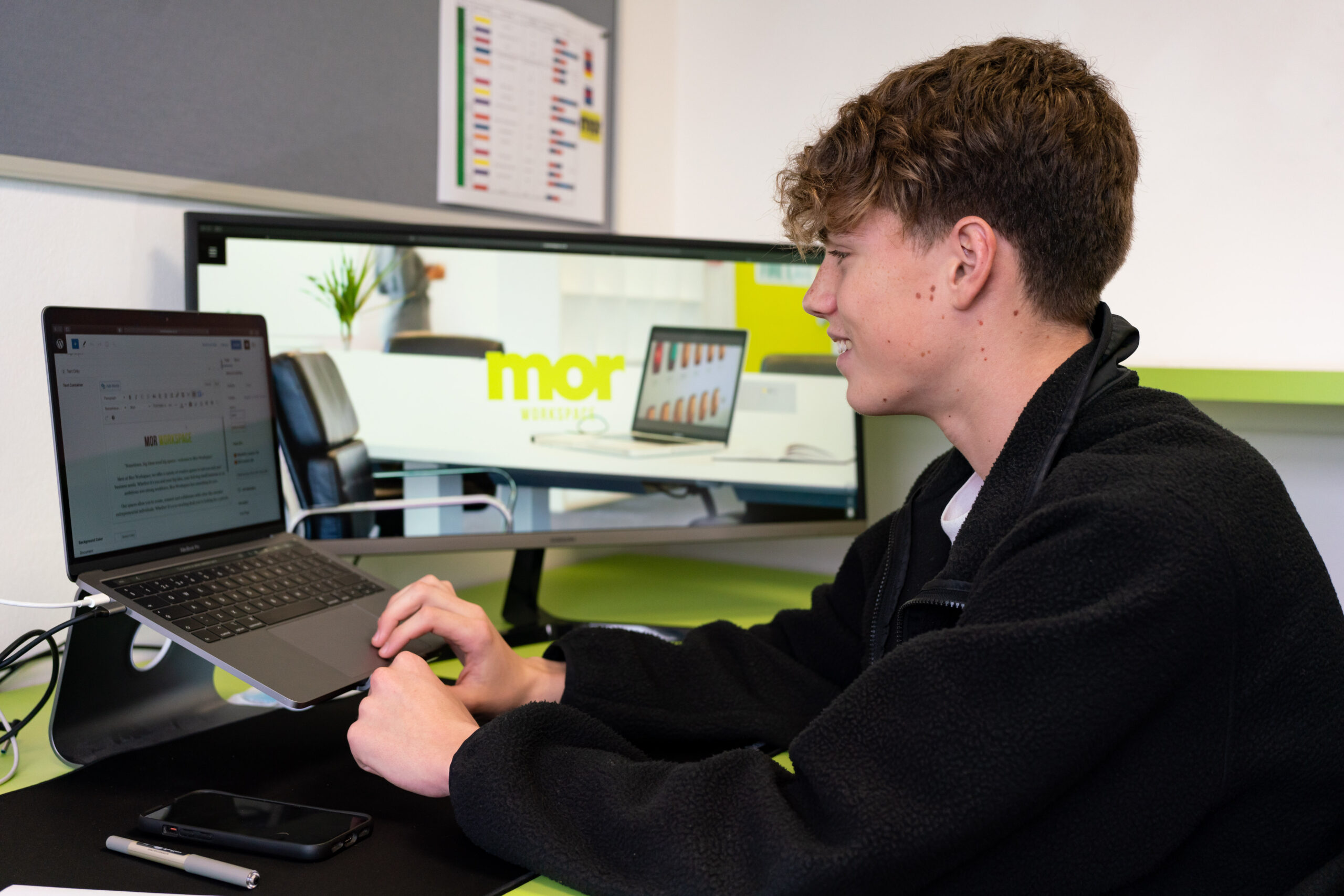 A young boy sits at a desk using a laptop while to the side another larger screen shows a company's website.