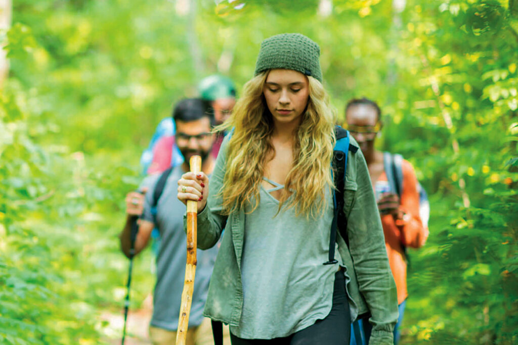 A young caucasian woman leads her friends on a hike through the woods. The sun glistens off the trees. They are looking down at the ground wearing backpacks and holding walking sticks.