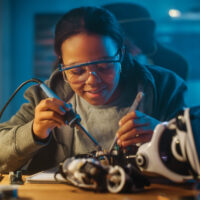 A young girl wearing goggles works on a piece of electrical equipment.