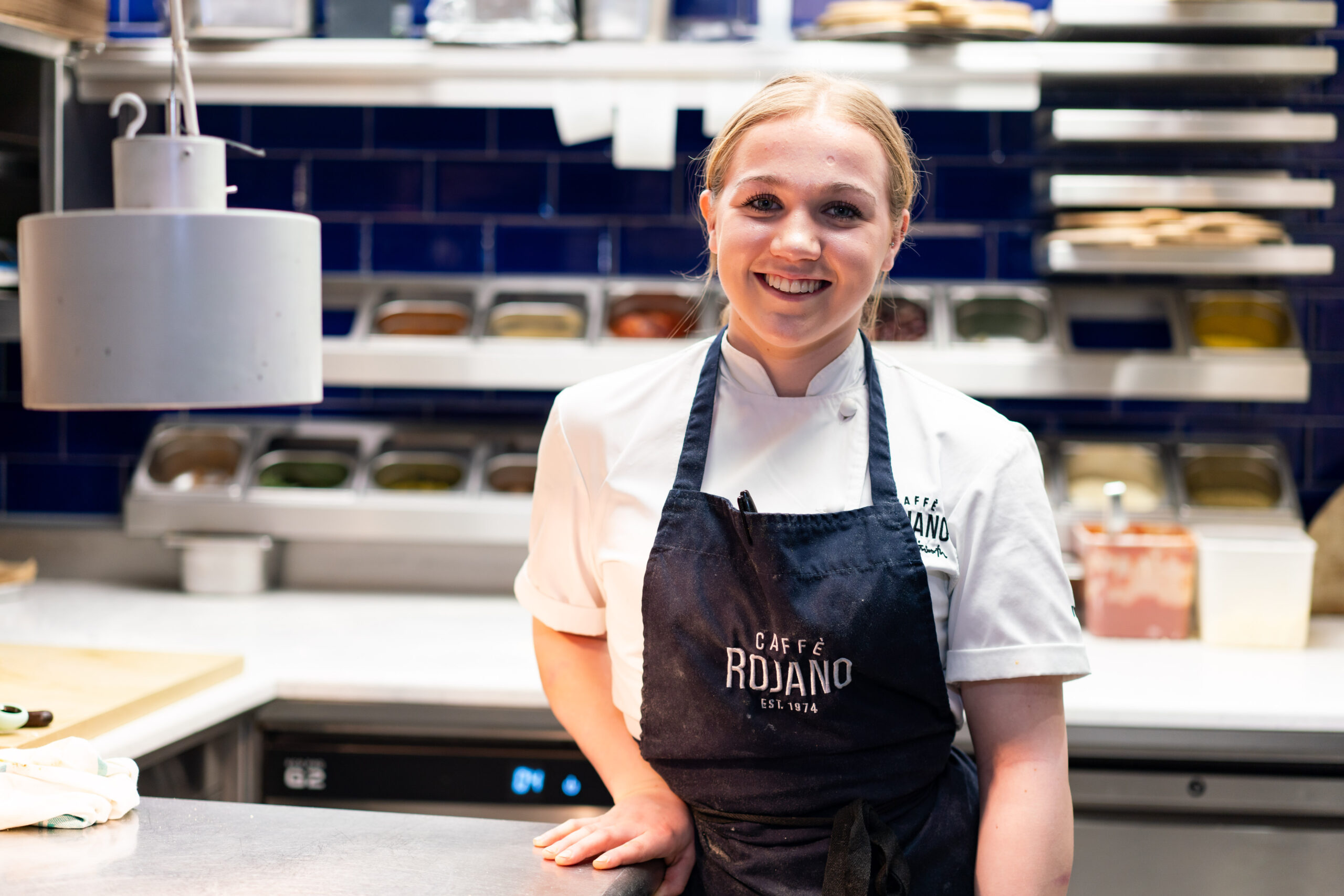 A young girl smiles at the camera wearing an apron in a professional kitchen environment.