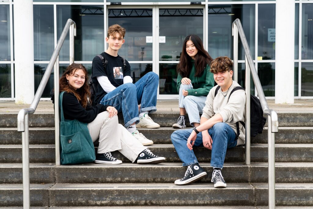 Four students sit on the steps outside of a College building. They look at the camera and smile.