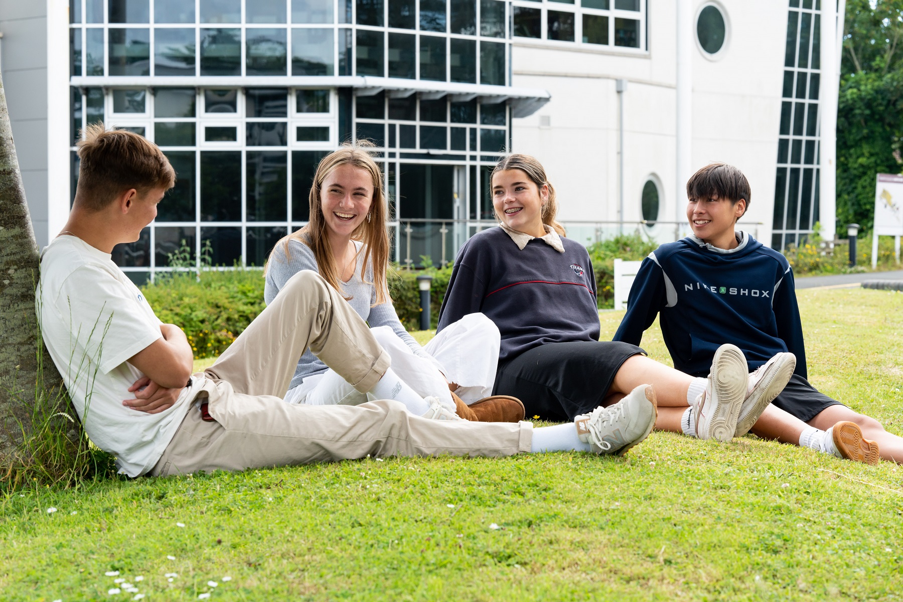 Two male and two female students sit on the grass outside of a college building. They are smiling and laughing.