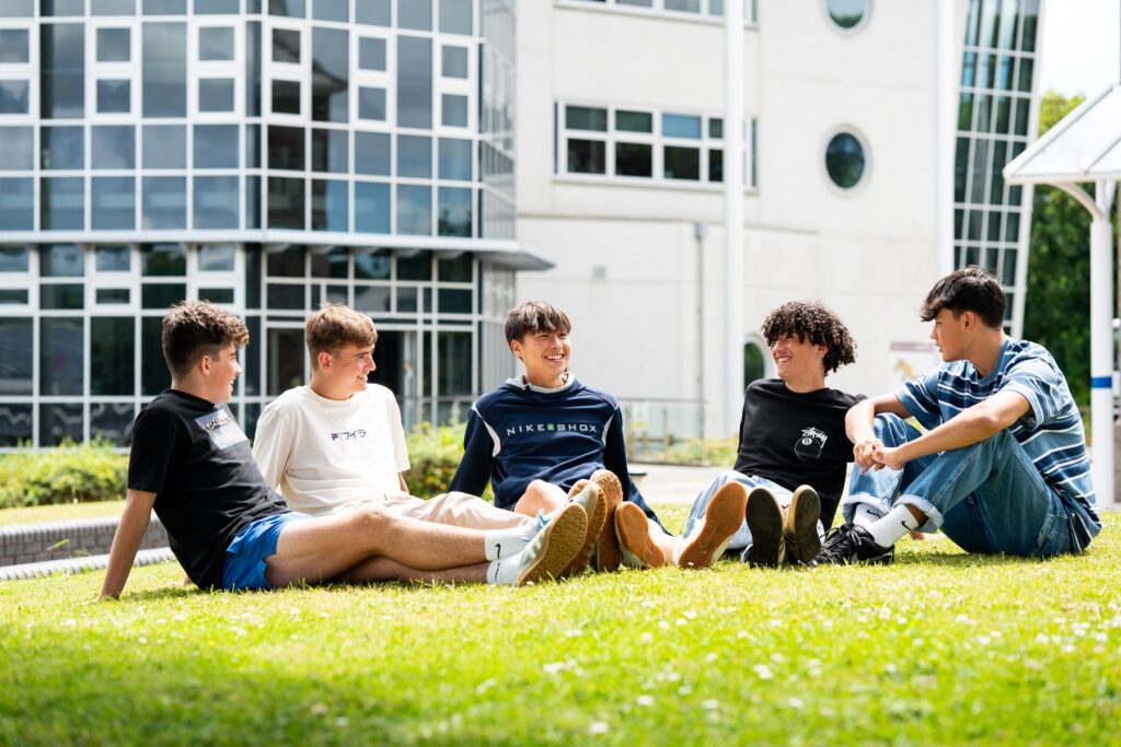 Five male students sit on the grass in front of a college building. They are smiling and laughing.