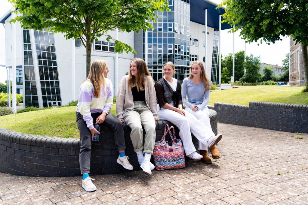 Four female students sit on a low wall in front of a college building. They are smiling at each other.
