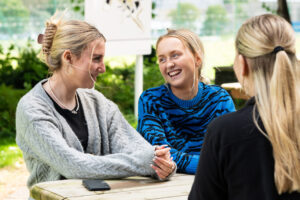 Three female students sit at a picnic bench, smiling and laughing. There is a mobile phone face down on the table.