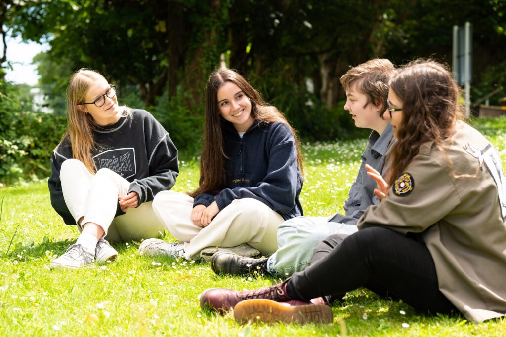 4 students sit on the grass in the sunshine. They are chatting.