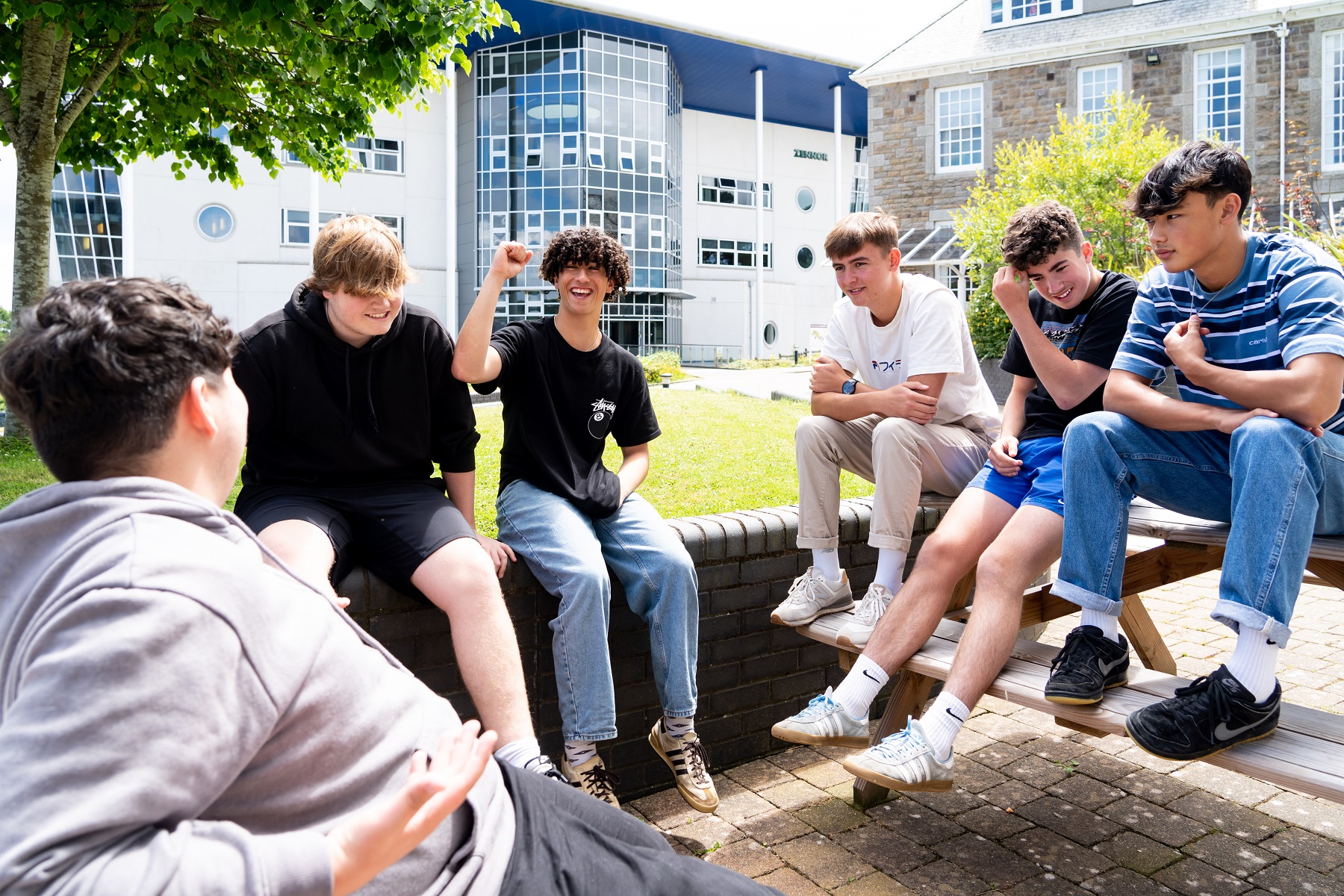 A group of male students sit on a low wall and picnic benches. They are chatting and laughing.