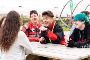 A group of friends are gathered around a picnic bench outside and smiling.