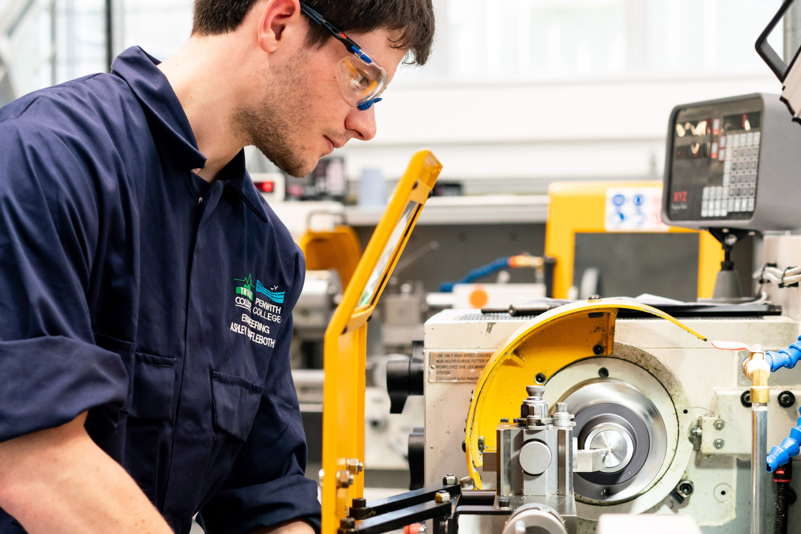 A student wearing overalls and protective eyewear looks down at engineering equipment.