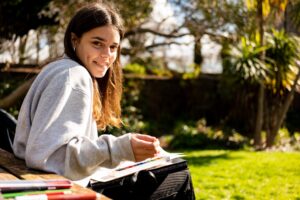 A student sat outside in the sun holds a paintbrush and has watercolour paints on her lap.
