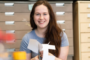 A students looks at the camera smiling. She is cutting a piece of paper with a pair of scissors.