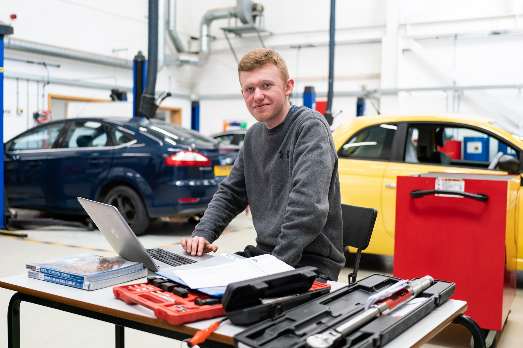 An automotive student works on a laptop in an automotive bay. There are cars in the background.