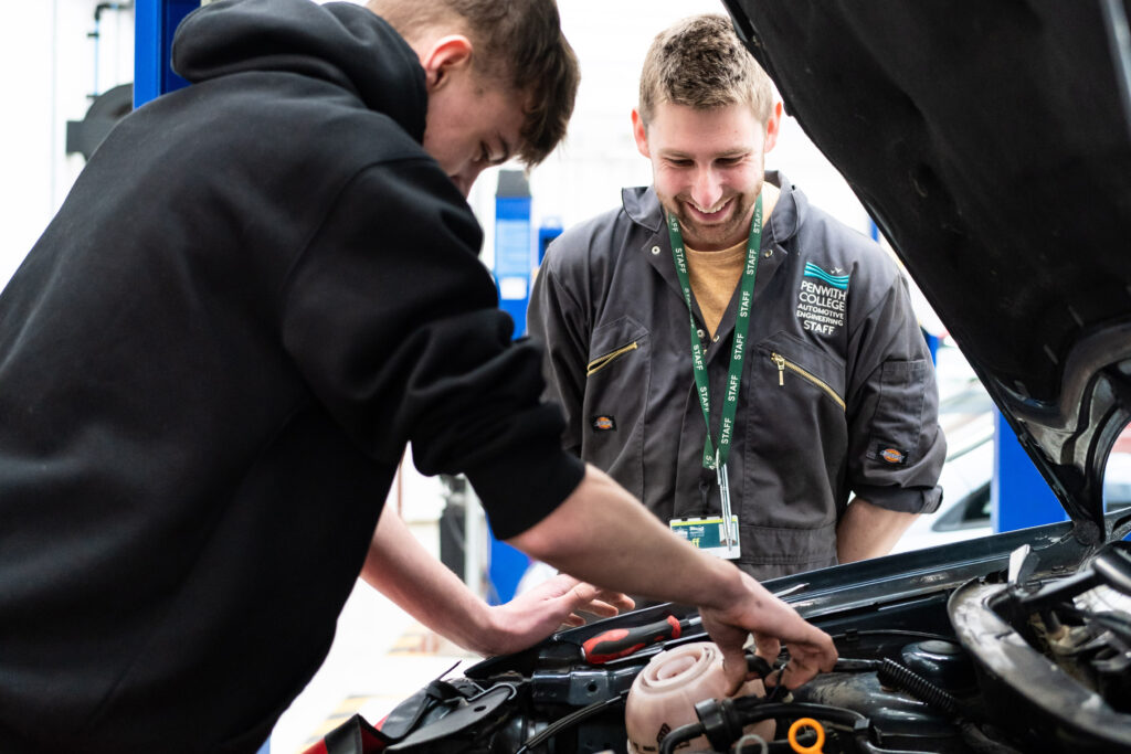 A tutor from the automotive department watches over a student as he works underneath the bonnet of a car.