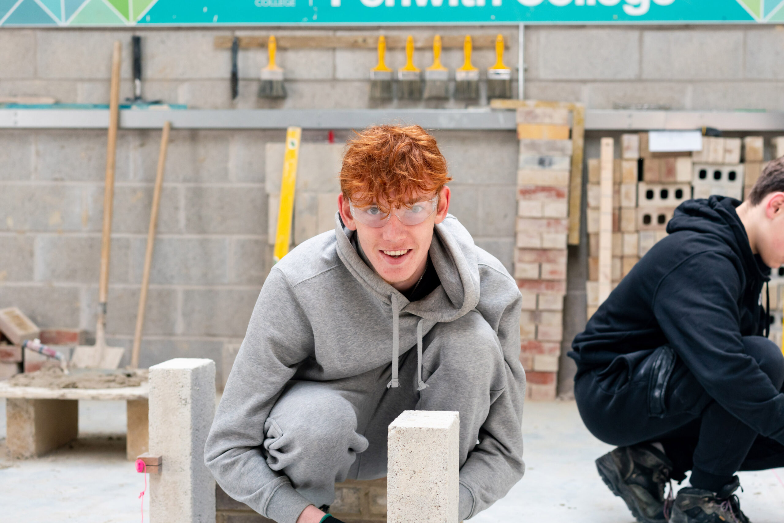 A bricklaying student in a grey hoodie smiles down the camera which laying blocks in one of the College's Construction areas.