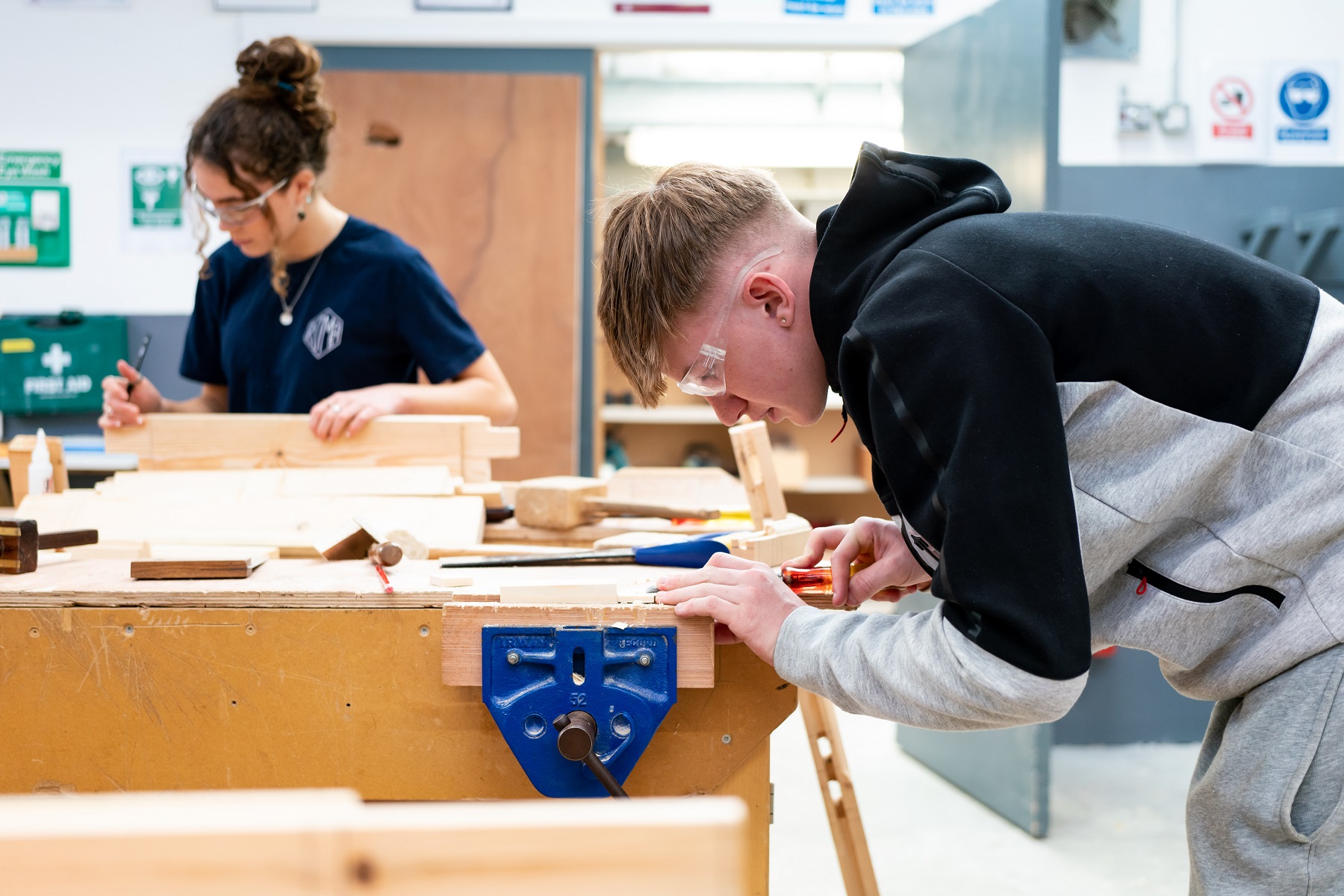 Two students are doing woodwork in the college. They both are wearing goggles and are focussed on their work.